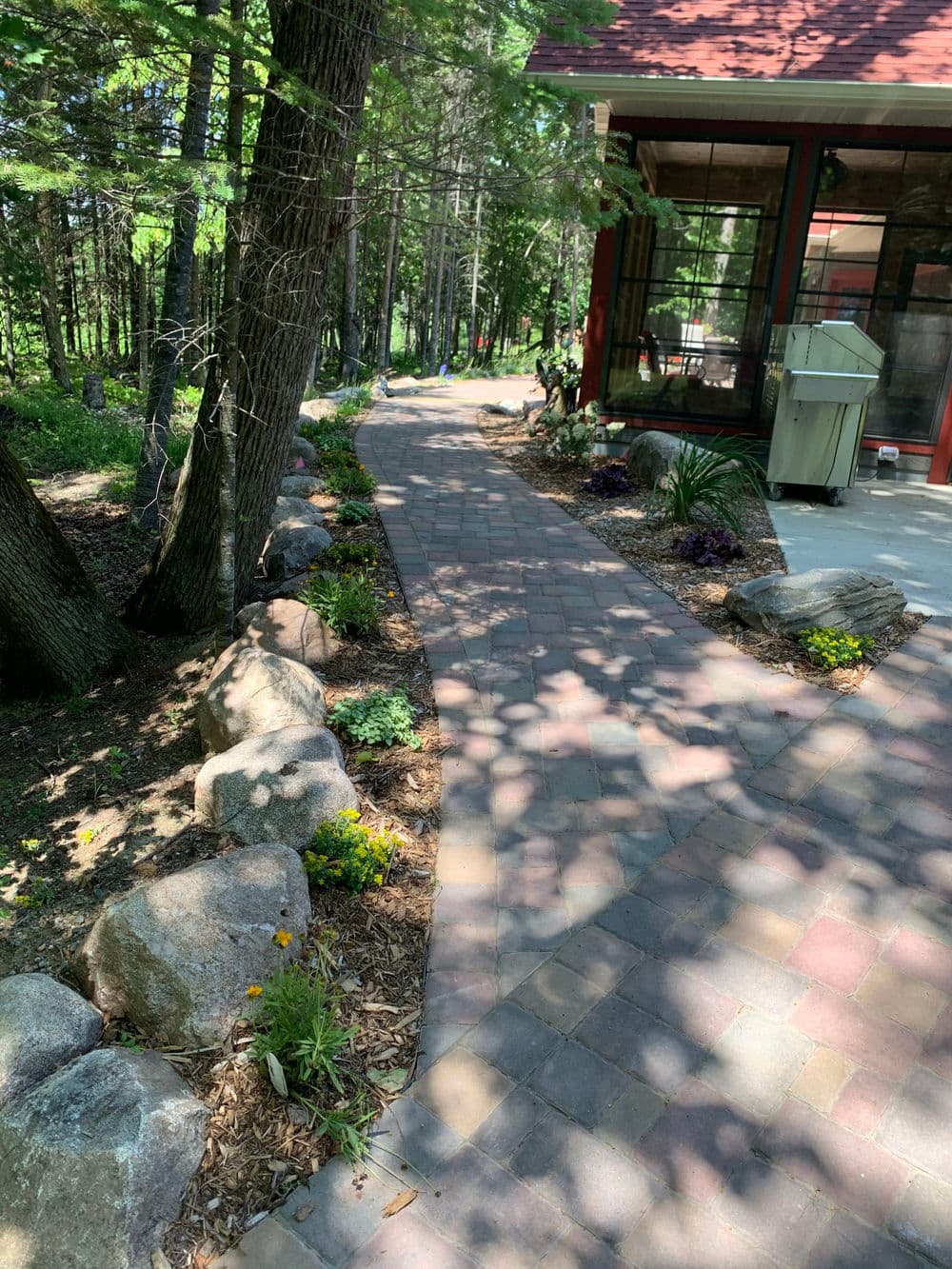 Curved stone pathway lined with flowers and rocks in a wooded garden setting.