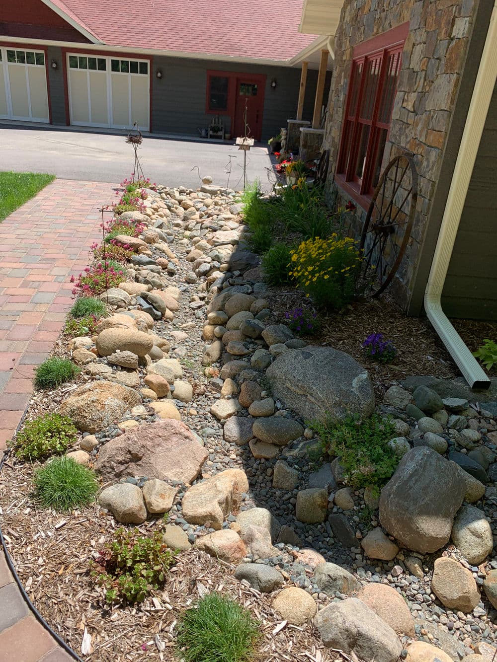Dry creek bed landscaping featuring stones, colorful flowers, and green plants beside a home.