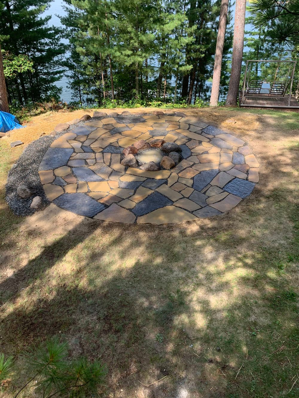 Natural stone fire pit area surrounded by trees near a lake, ideal for outdoor gatherings.
