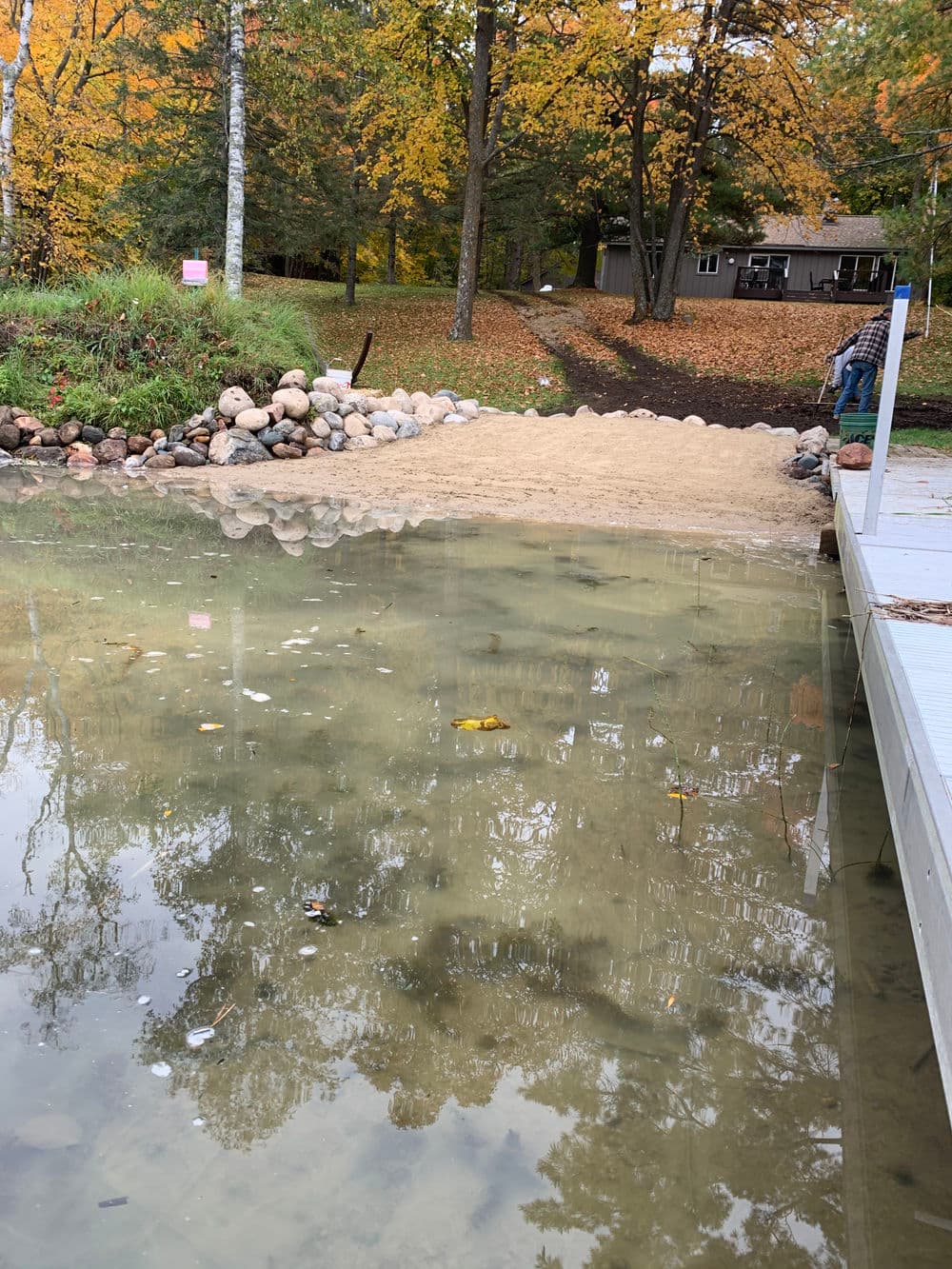 Sand path leading to a reflective lake surrounded by autumn trees and a cabin.