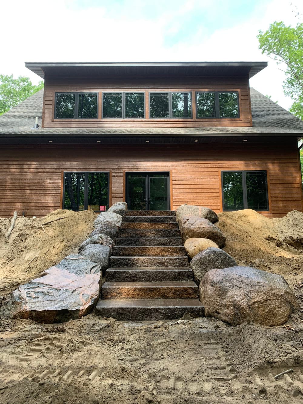 Modern wooden house with large windows and stone steps leading to the entrance, surrounded by landscaping.