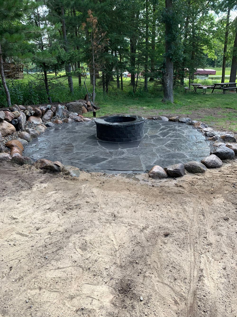 Stone fire pit surrounded by a circular stone patio in a wooded outdoor setting.