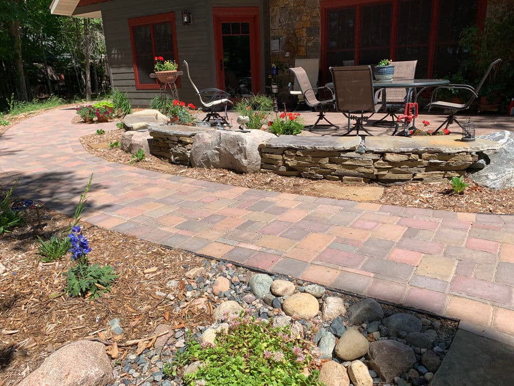 Residential garden pathway with flower beds, stone seating area, and patio furniture.