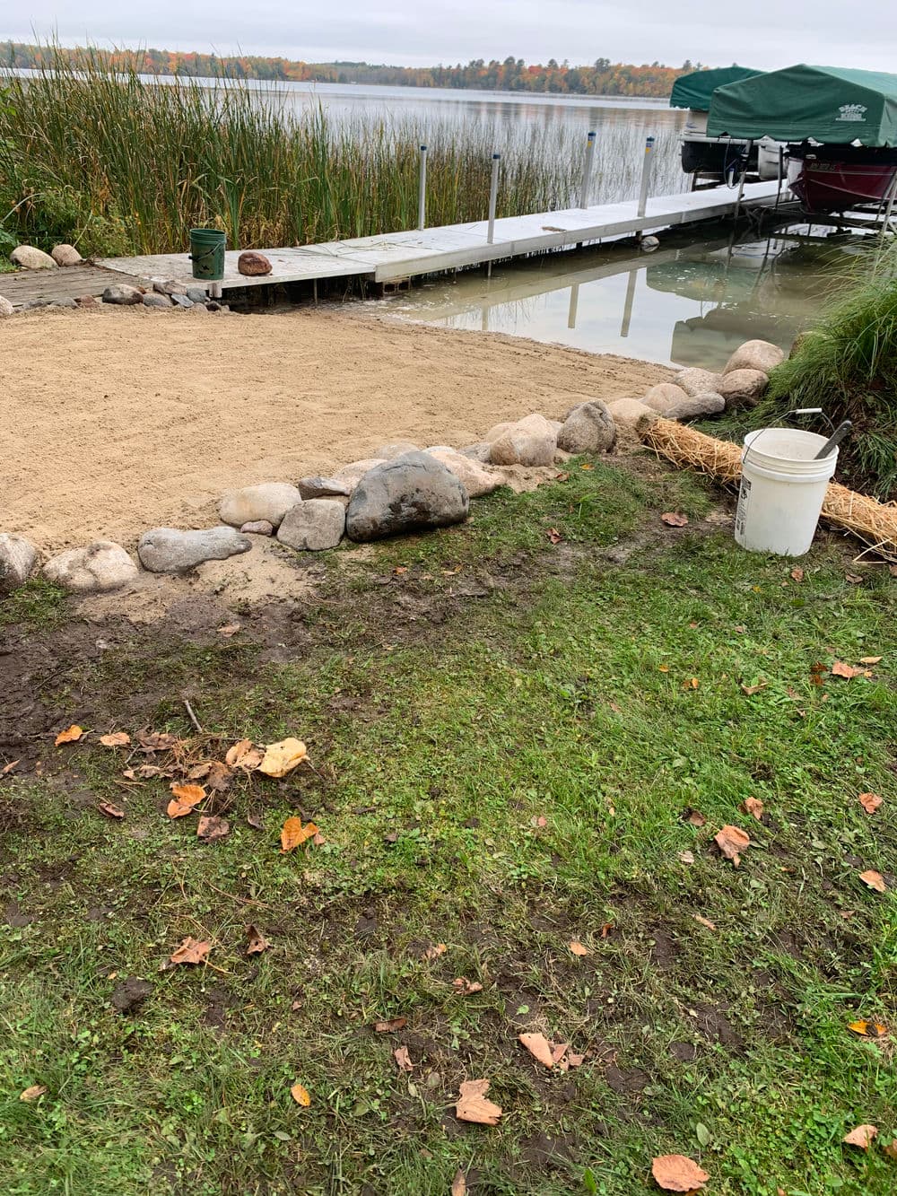 Lakefront scene with sandy area, dock, and boat, surrounded by grass and autumn foliage.