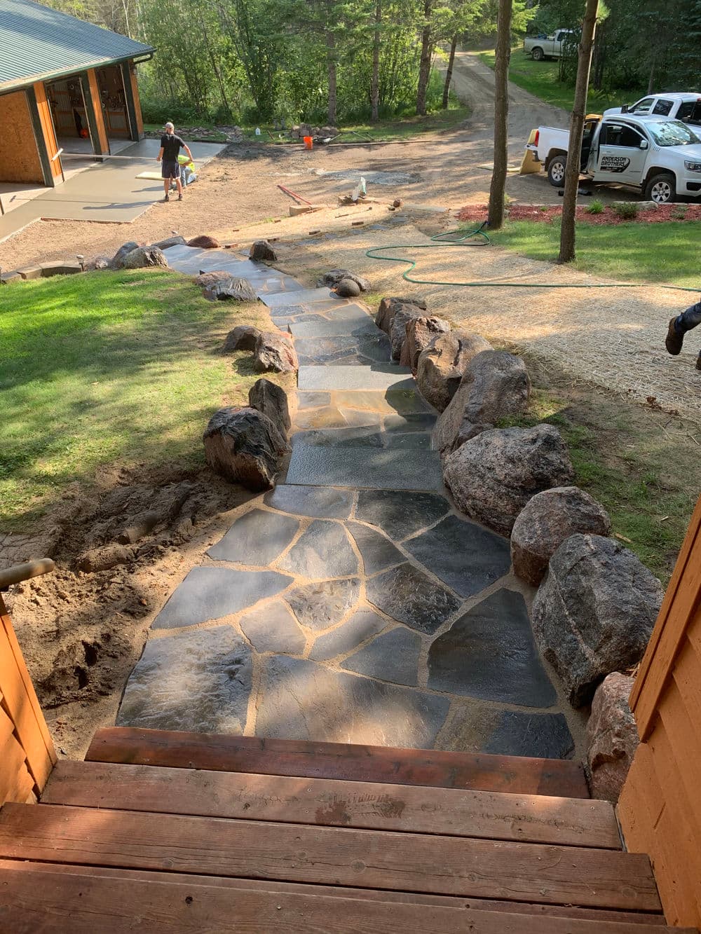 Stone path bordered by rocks leading through a landscaped yard with a driveway in view.
