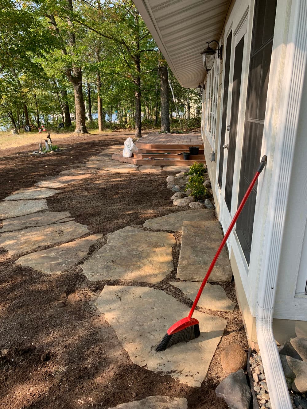 Garden pathway with stone pavers, broom, and wooden deck in a wooded outdoor setting.
