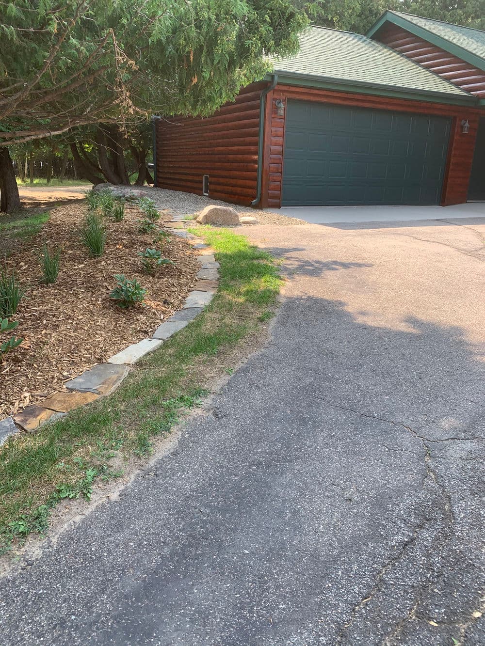 Log cabin garage with landscaped path, greenery, and stone edging in a tranquil outdoor setting.