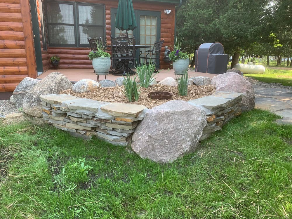 Landscaped stone garden bed with greenery near a wooden cabin and outdoor seating area.
