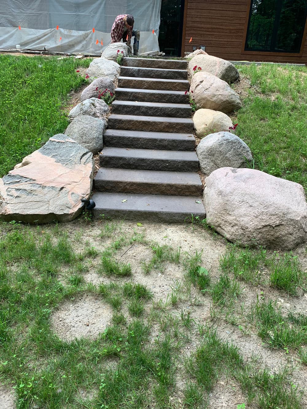 Stone steps leading to a house, bordered by large rocks and blooming flowers in a grassy area.
