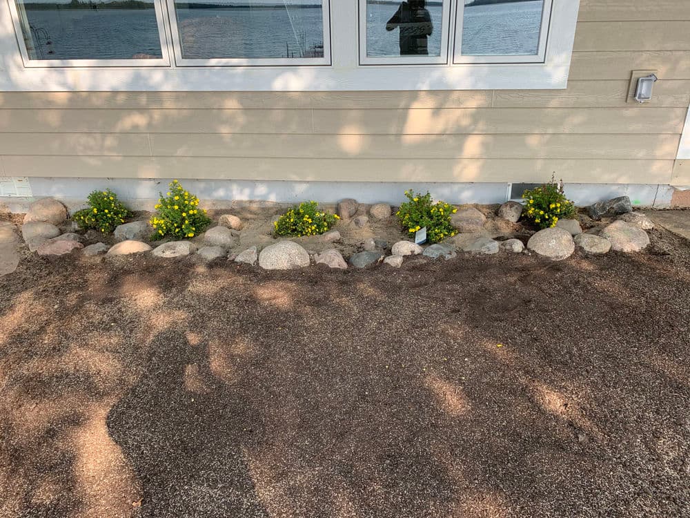 Flower bed with yellow flowers and rocks by a lakeside home foundation.