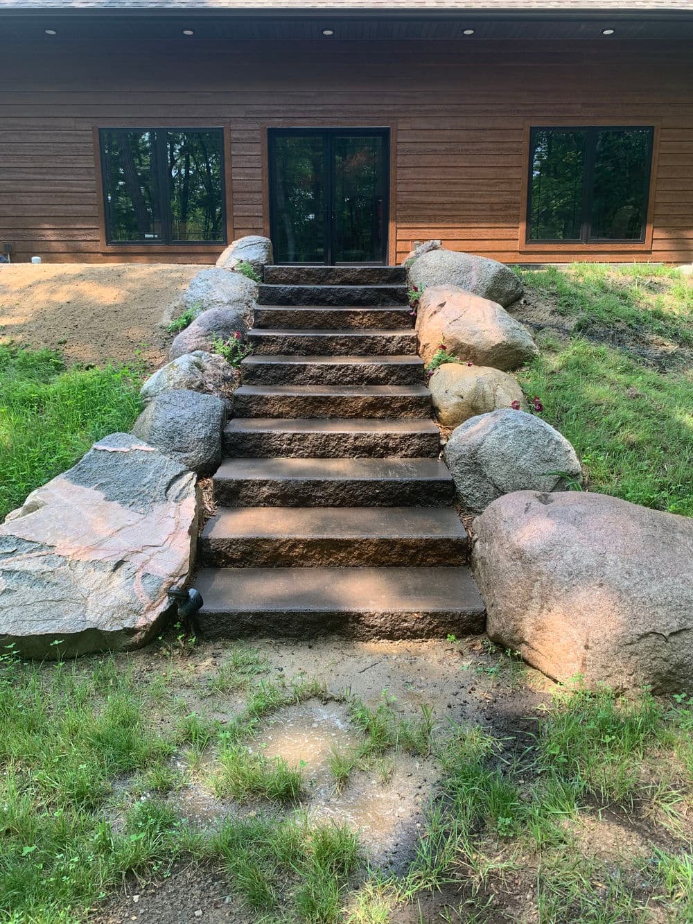 Concrete steps adorned with large stones lead to a modern wooden house in a green landscape.