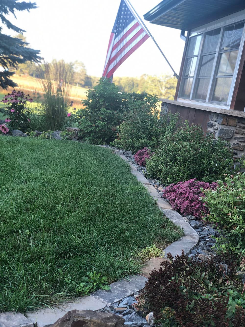 Lush green lawn with blooming flowers and an American flag near a cozy house.