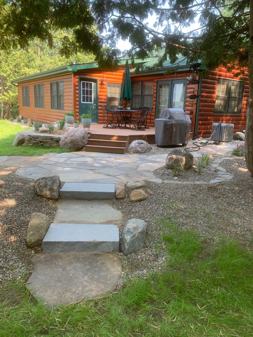 Log cabin with a stone pathway, patio furniture, and greenery in a tranquil outdoor setting.