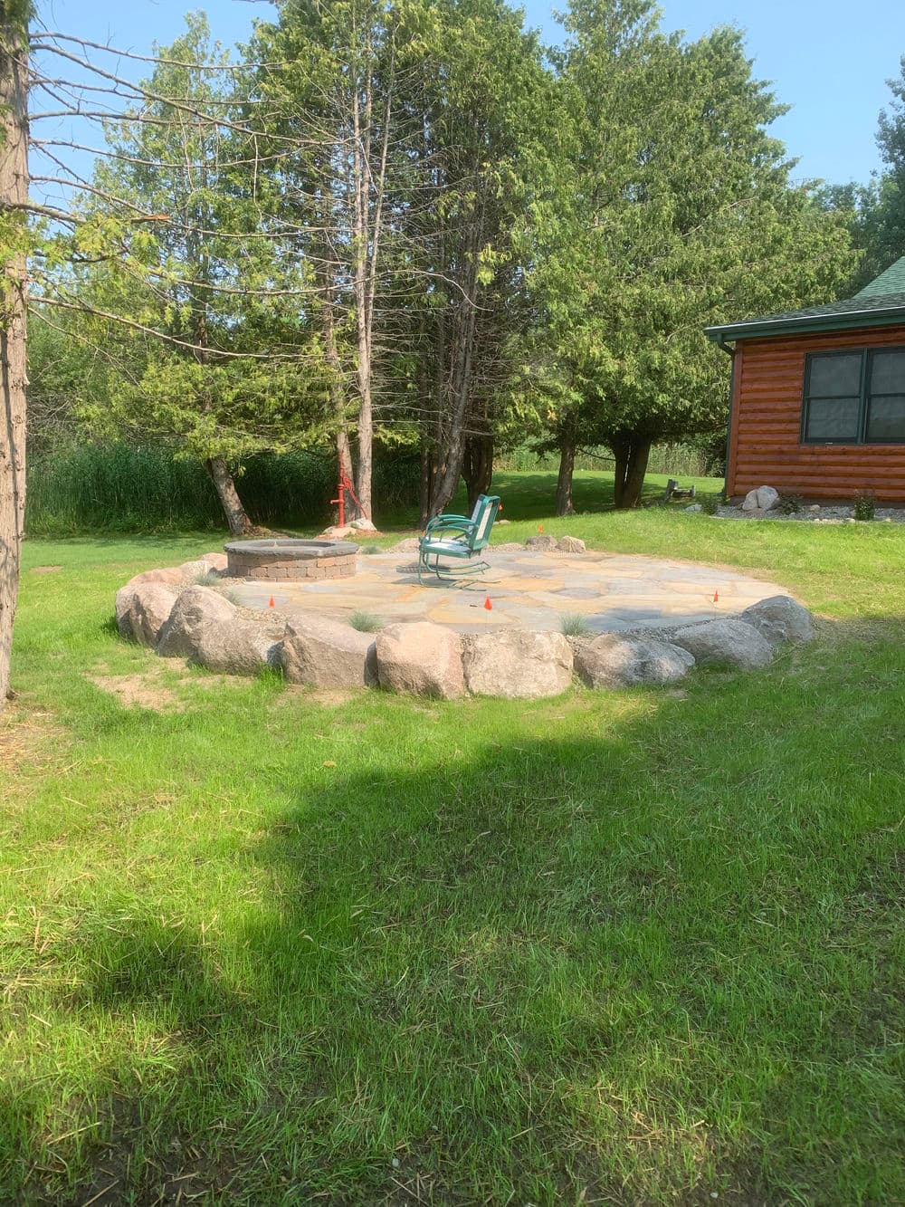 Lawn with stone patio and single green chair surrounded by trees near a cabin.