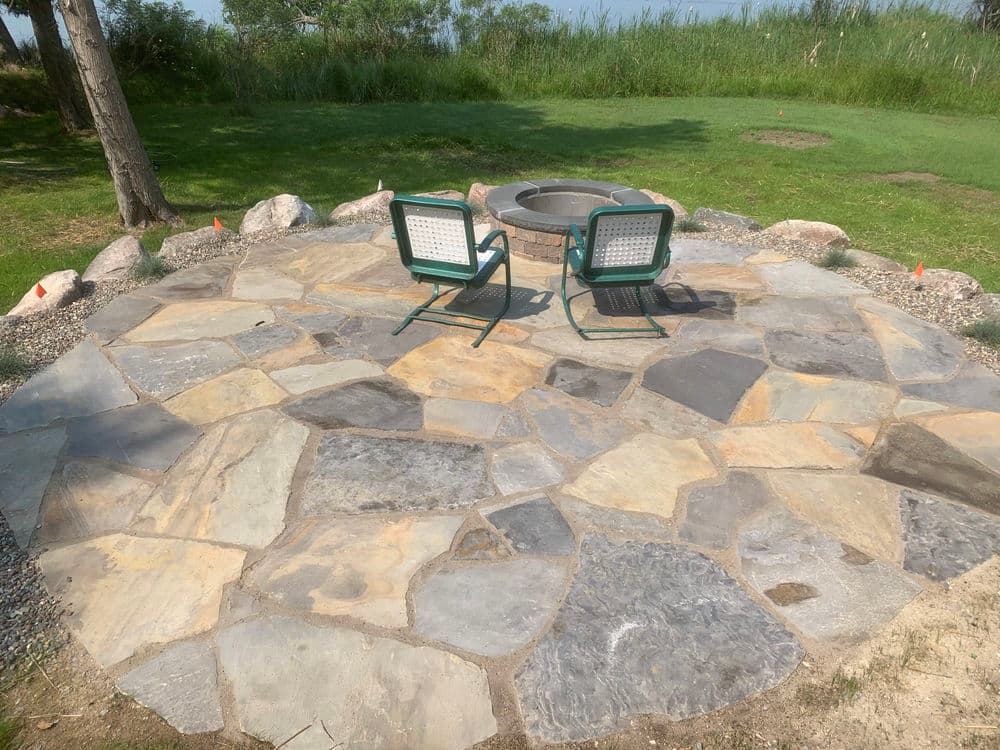 Stone patio with fire pit and two green chairs in a serene outdoor setting.