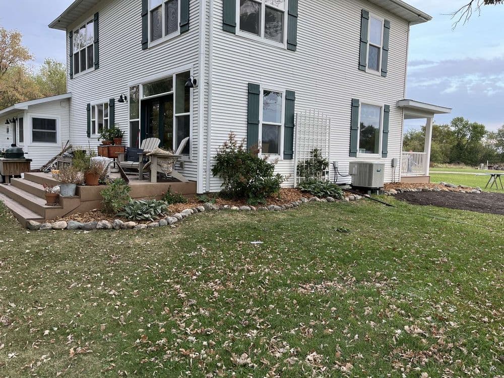 Two-story white house with green shutters, garden, and patio furniture in a grassy yard.