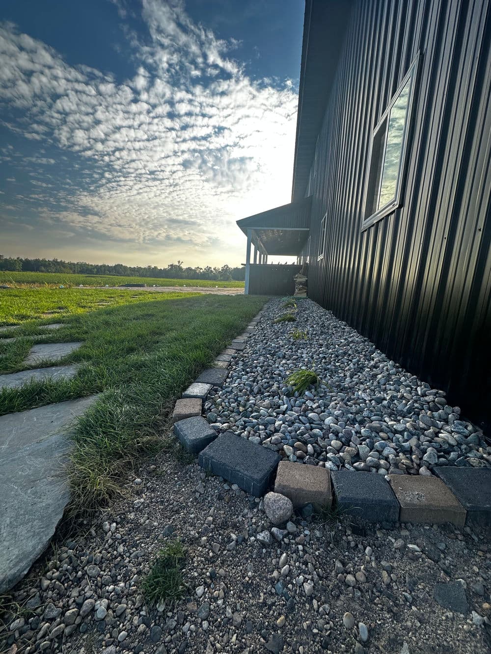 Black metal building beside a rock-lined path and grassy field under a cloudy sky.