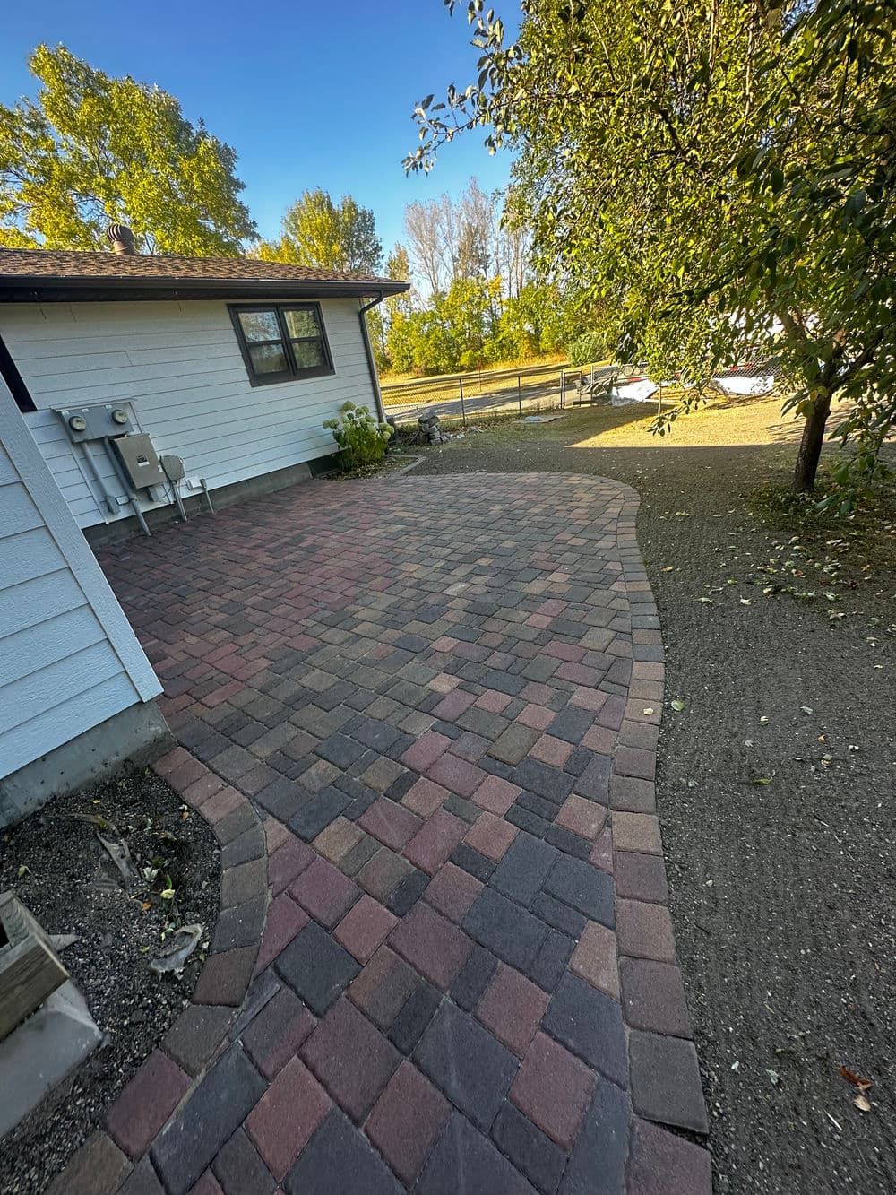 Paved patio with brick pattern beside a house, surrounded by trees and open space.