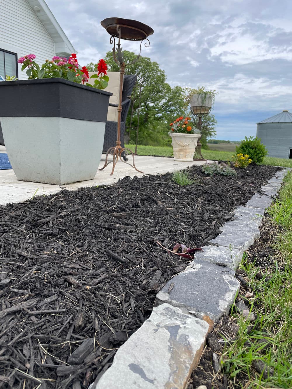 Flower garden with decorative planters, mulch, and a stone border under cloudy sky.