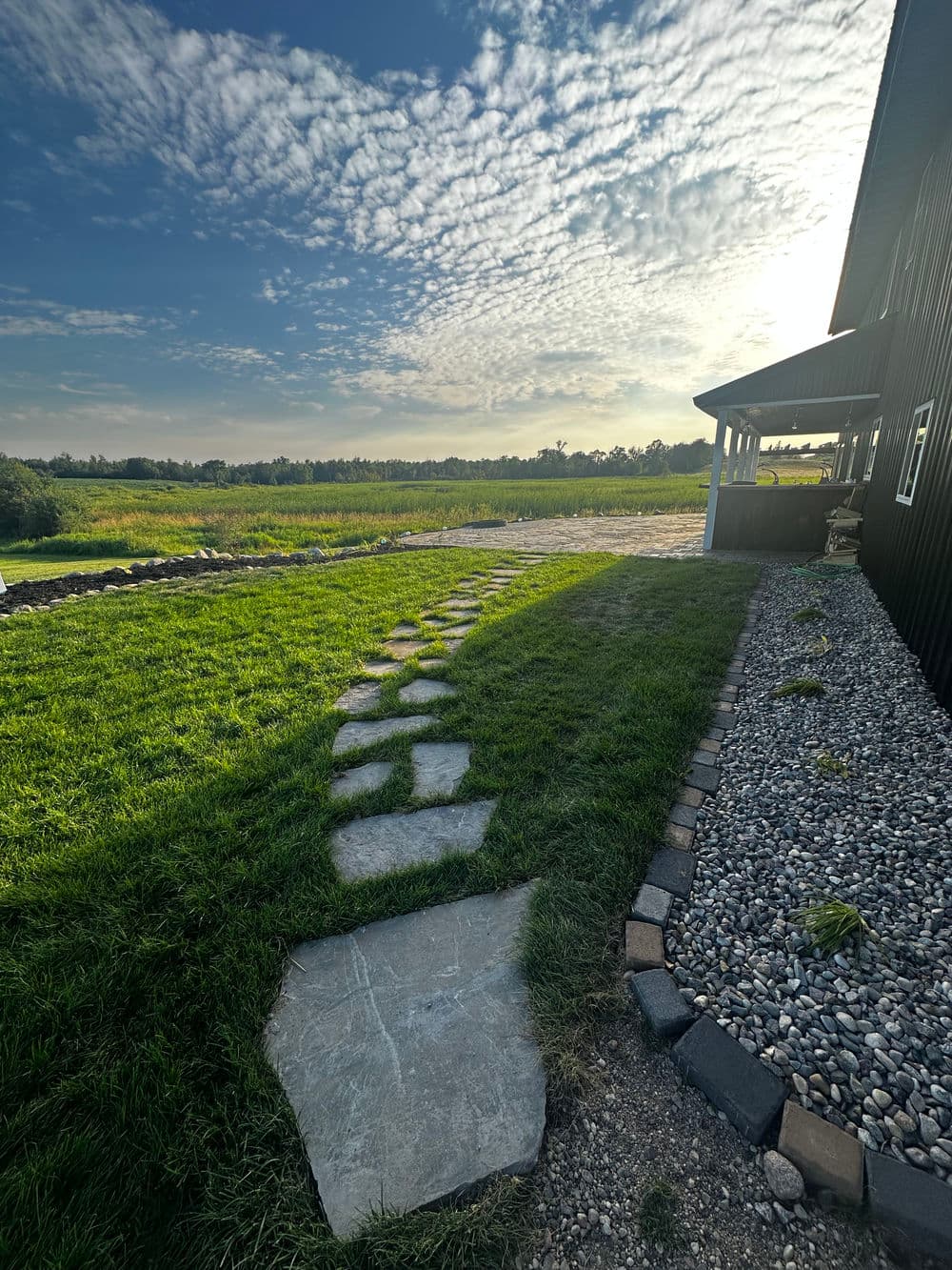 Sunlit stone path leading to a vibrant green field under a blue sky with clouds.