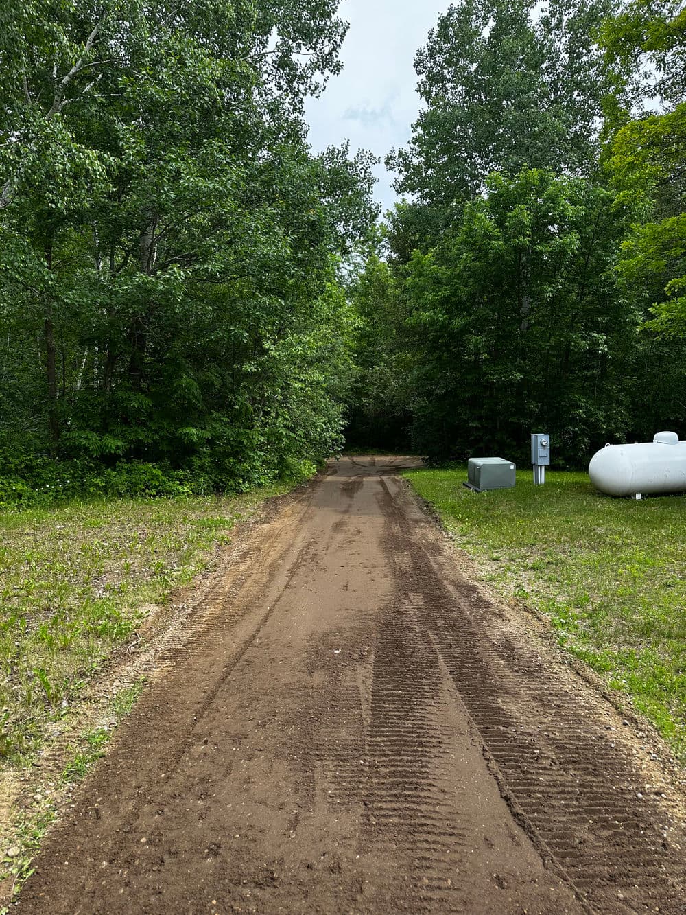 Dirt path leading through lush green trees with a propane tank nearby.
