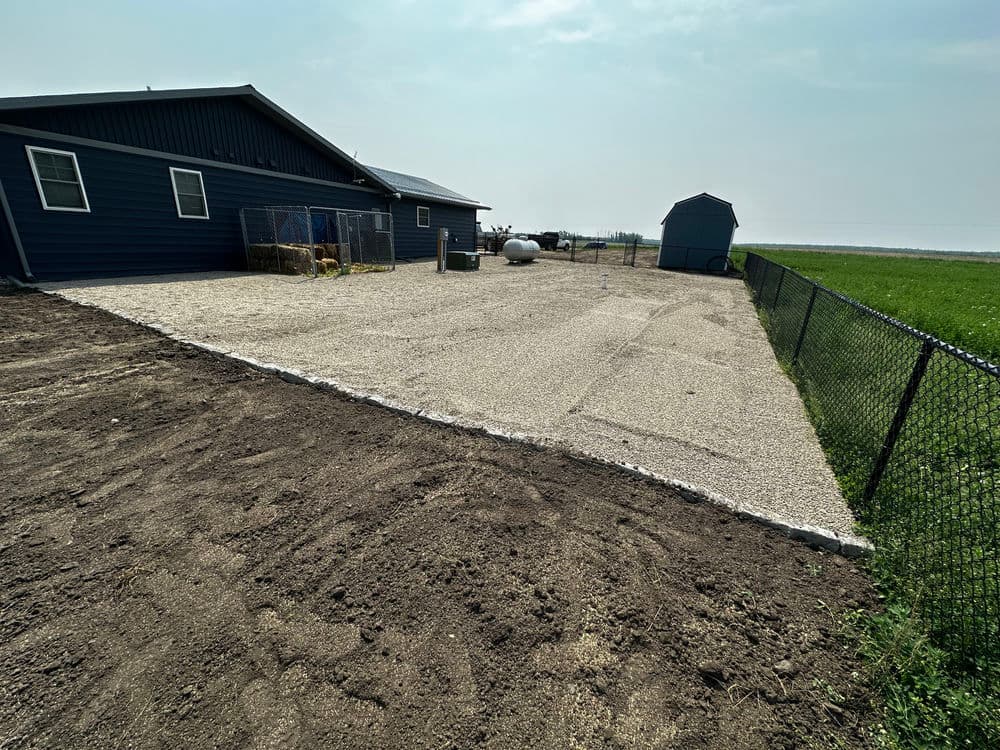 Gravel patio area beside a blue house, with a fenced yard and shed in the background.