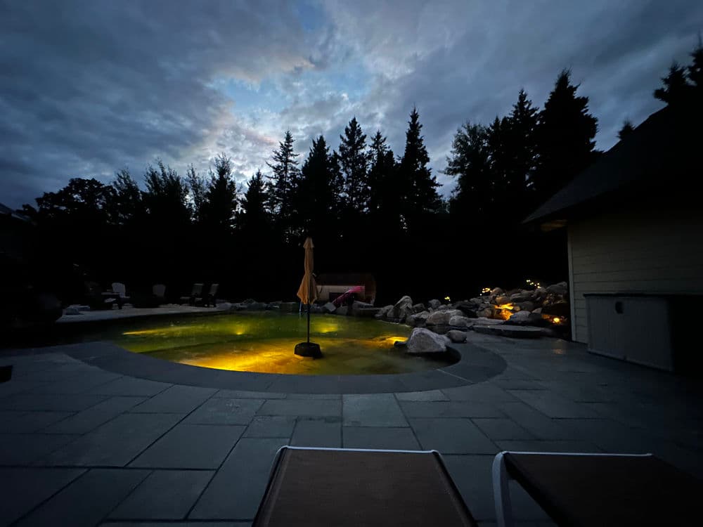 Serene nighttime pool setting with illuminated water, surrounded by trees and stone patio.