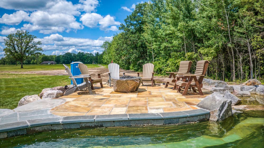 Cozy outdoor seating area with stone fire pit surrounded by lush greenery and blue sky.