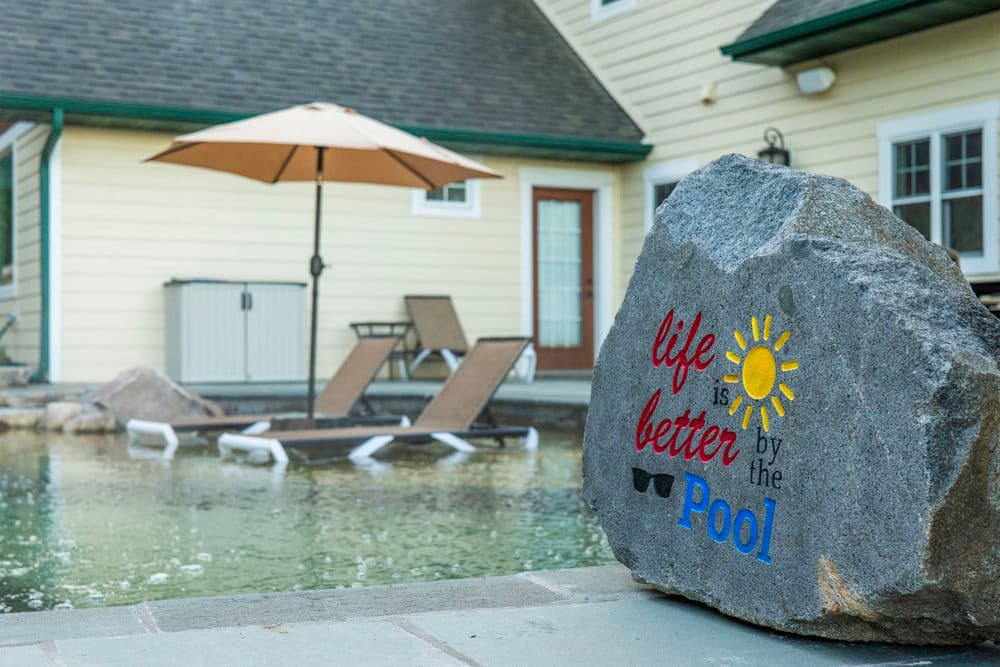 Stone with 'Life is better by the pool' near a relaxing pool area with lounge chairs and umbrella.