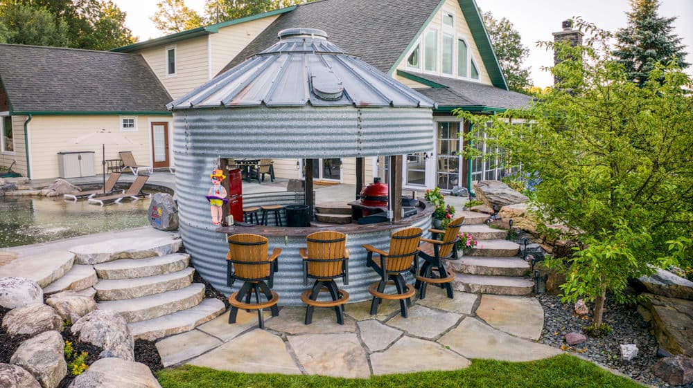 Outdoor bar made from a metal silo with stone steps and seating in a backyard setting.