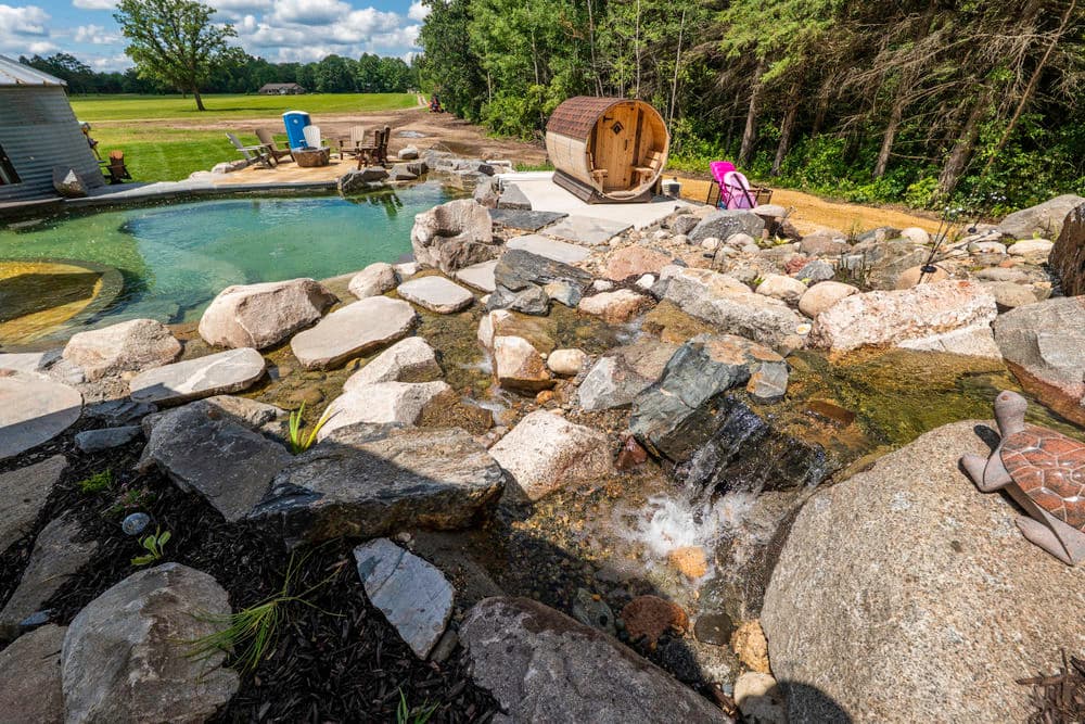 Scenic backyard with a pool, natural stone waterfall, and barrel sauna amid greenery.