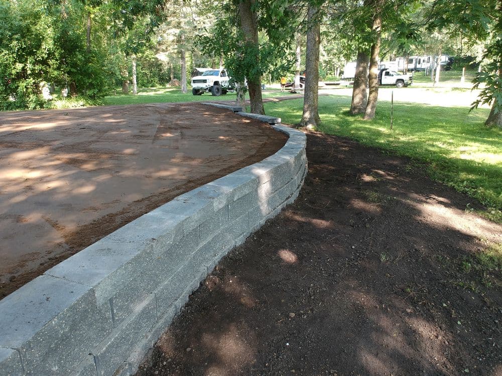 Stone retaining wall construction in a shaded outdoor area with vehicles nearby.