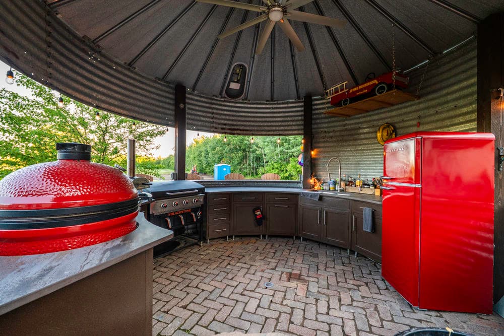 Outdoor kitchen with red refrigerator, grill, and covered seating area amid greenery.
