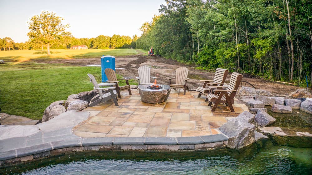 Outdoor fire pit area with wooden chairs, surrounded by greenery and a portable restroom nearby.