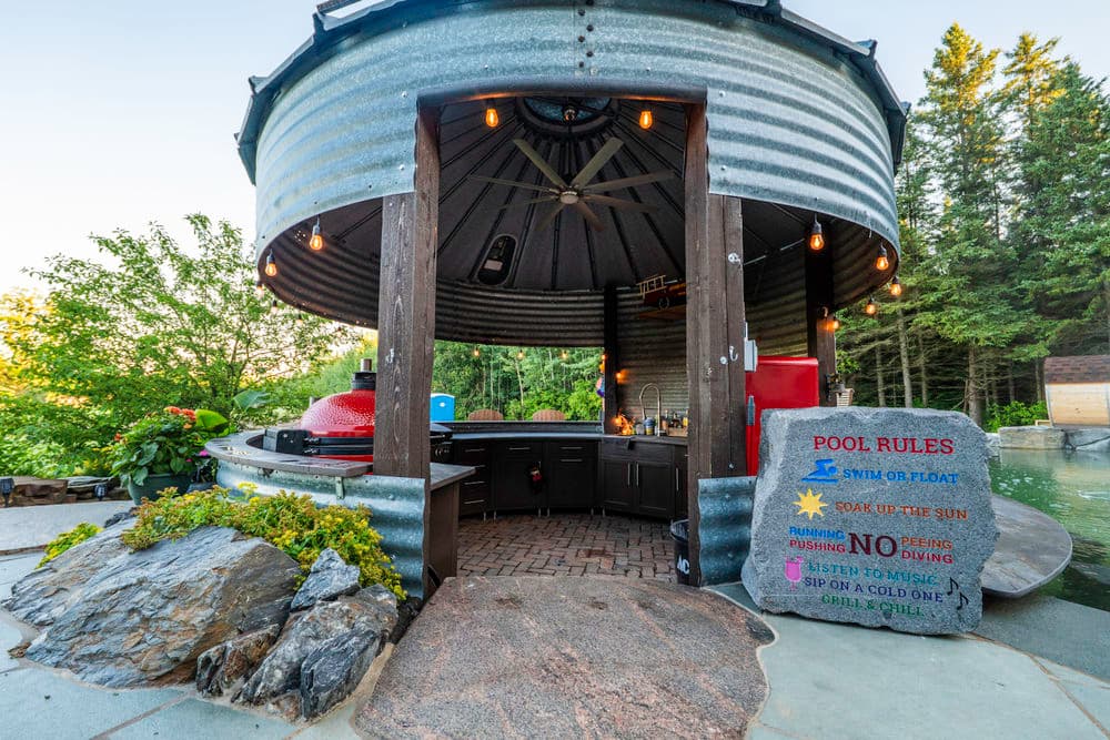 Outdoor kitchen with metal roof, surrounded by greenery, featuring grill and pool rules sign.