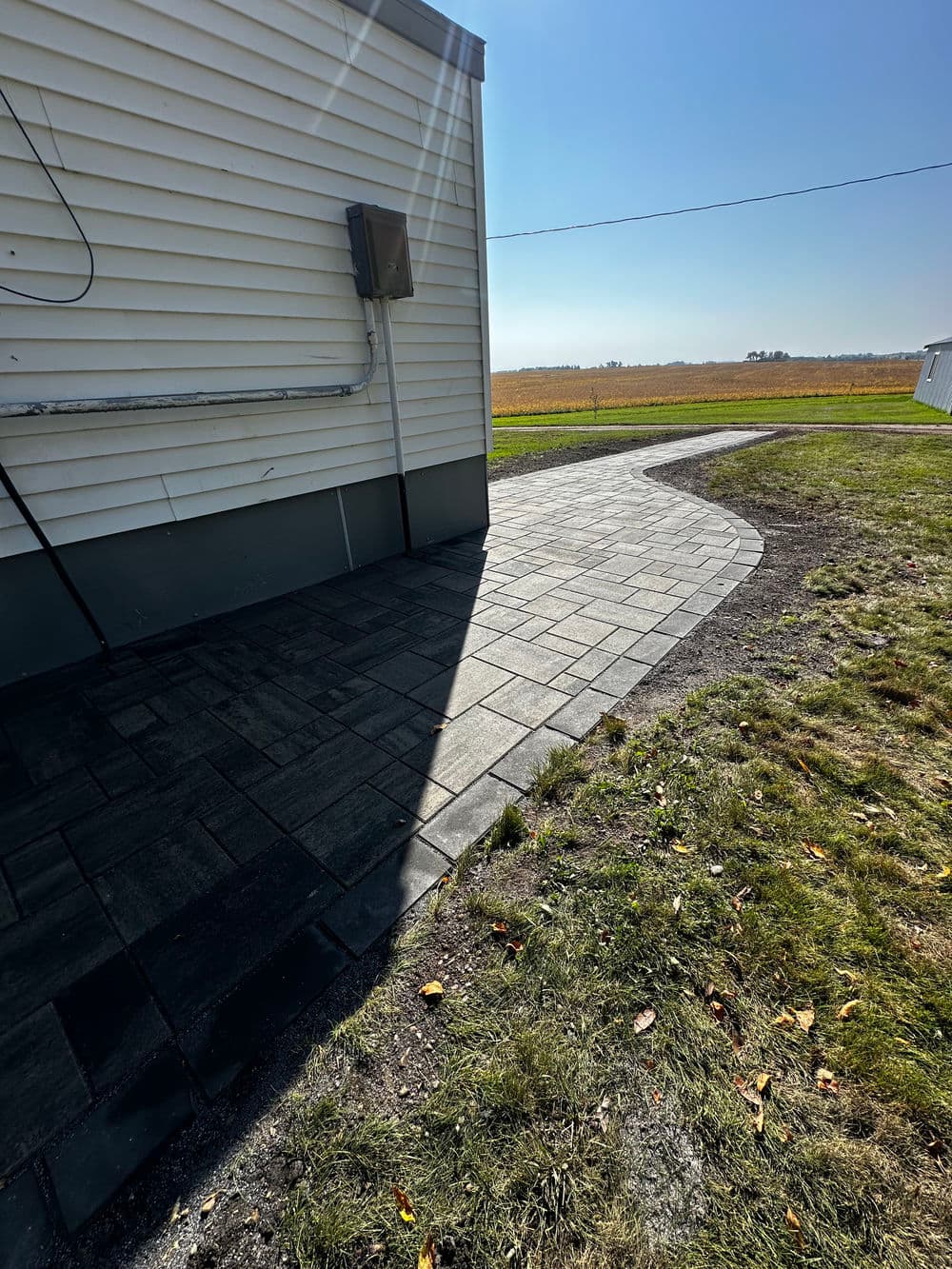 Paved walkway leading from house to open field, with clear blue sky and grass nearby.