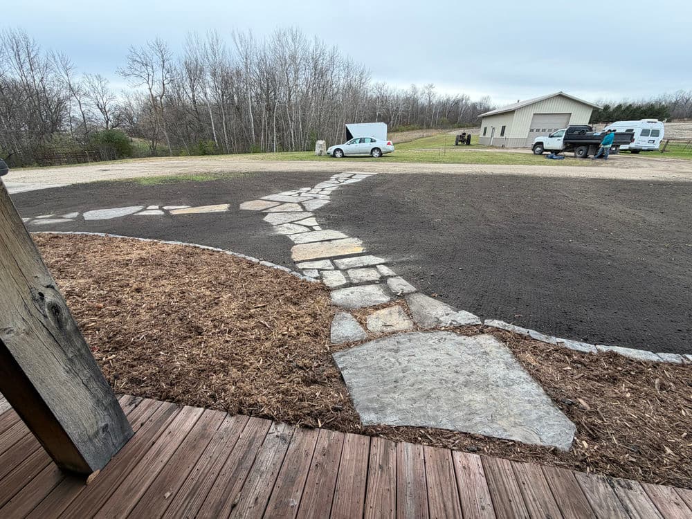 Stone pathway leading to a garage with vehicles, surrounded by bare soil and trees.
