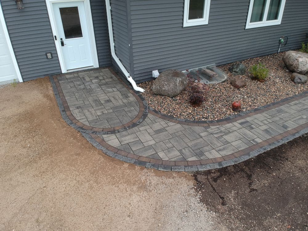Paved walkway with stone borders leading to a gray house entrance and landscaped garden.