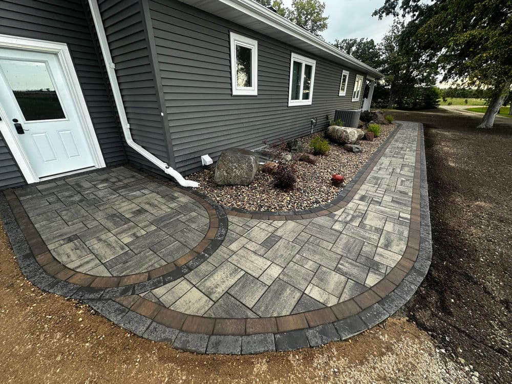 Paver pathway with curved design leading to a grey house exterior and landscaped garden.